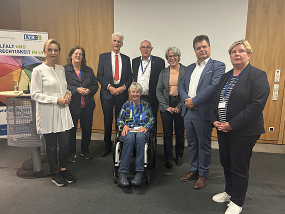 Gruppenfoto, 7 Teilnehmer*innen stehen im Halbkreis vor einer Wand mit Holztäfelung und weißer Projektionsfläche. In der Mitte des Halbkreises sitzt Sigrid Arnade im Rollstuhl. Links an der Seite ein farbiger Aufsteller des LVR sowie ein Stehtisch.