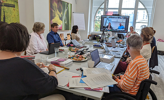 11 Personen rund um einen Tisch (2 davon per Videobildschirm). Auf dem Tisch Laptops, Papiere, Teller mit Süßigkeiten