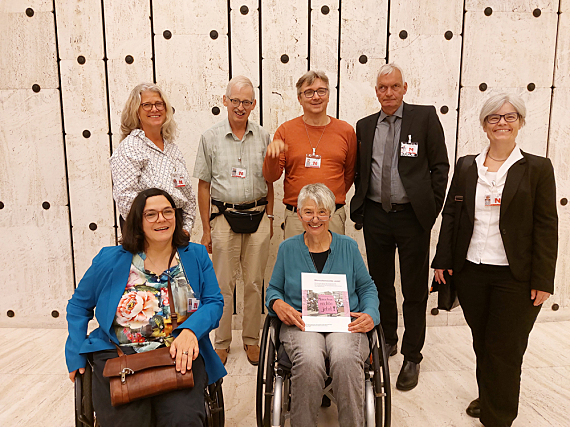 Foto der 7 Delegierten. Alle mit Namensschild. Sigrid Arnade hat den Paralellbericht auf dem Schoß, hält ihn in die Kamera. Im Hintergrund eine helle Wand aus senkrechten Segmenten mit Löchern