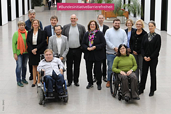 Gruppenbild der Bündnisinitiative Barrierefreiheit im BMAS. 12 Personen stehen in eienr lockeren Reihe, vor ihnen sitzen 2 Personen im Rollstuhl. Die Gruppe ist paritätisch zusammen gesetzt.